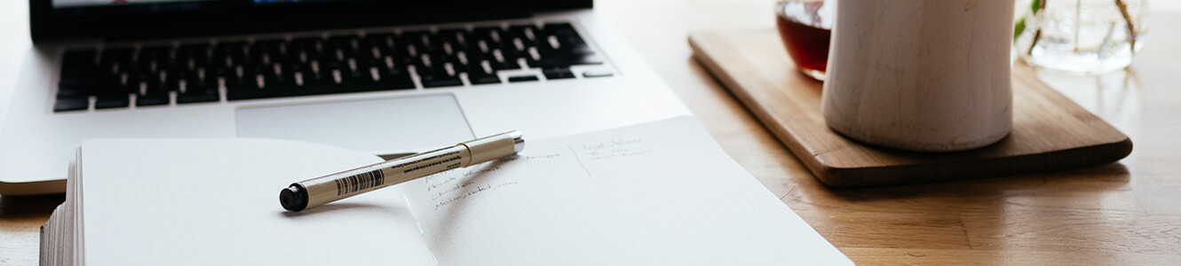 Photo of pen set on top of a journal in front of a laptop computer.